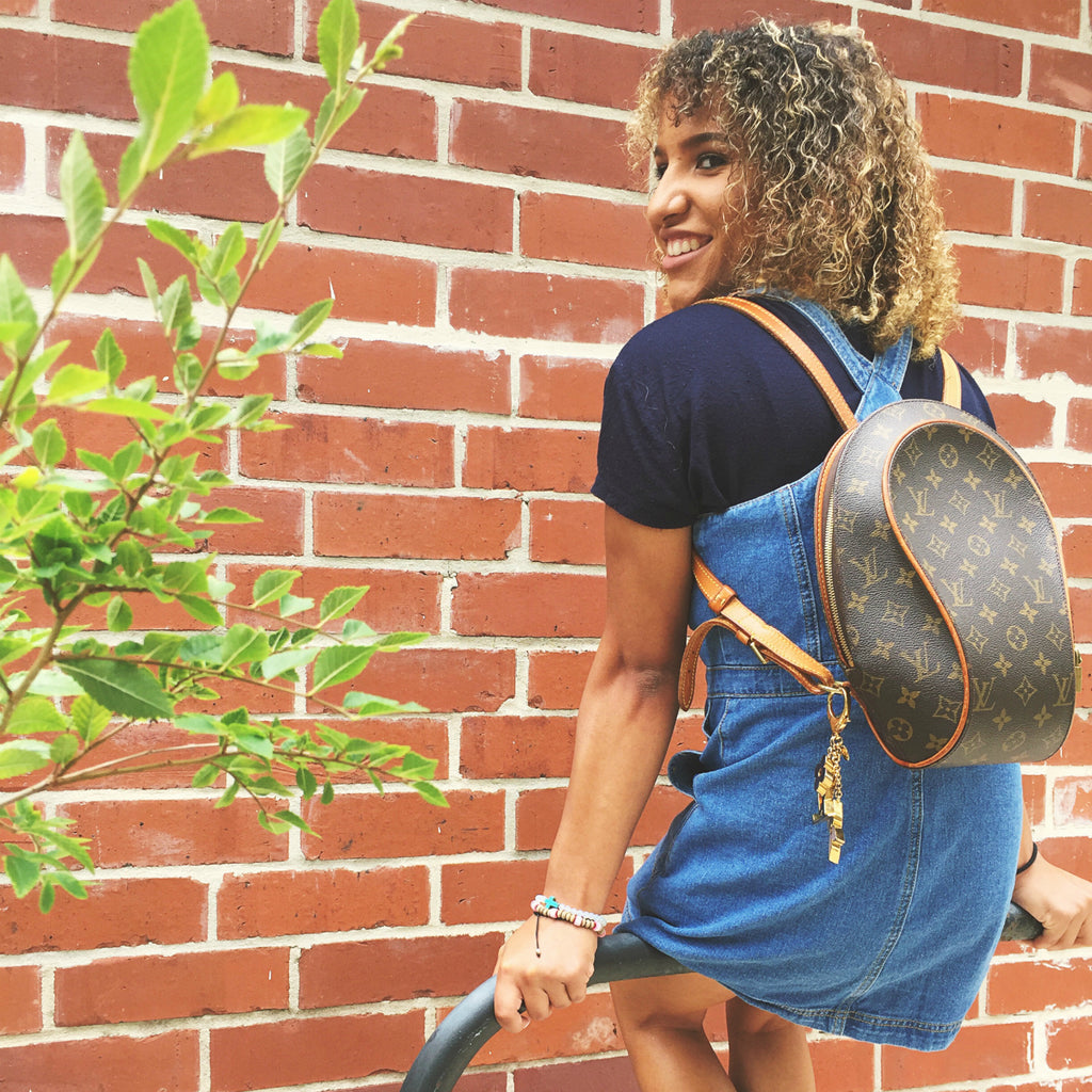 young woman wearing louis vuitton backpack denim dress next to brick wall